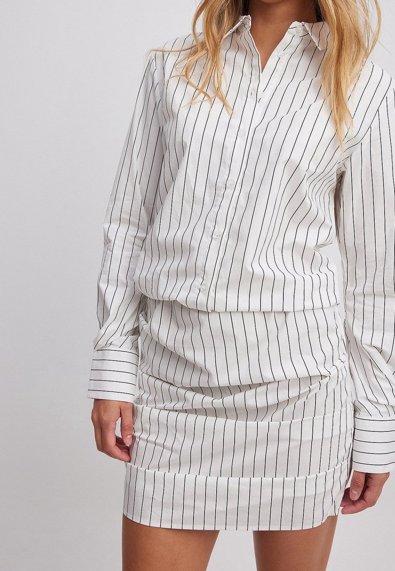 Femme portant une chemise blanche à manches longues boutonnée et une jupe blanche assortie avec de fines rayures verticales noires, debout devant un fond uni.