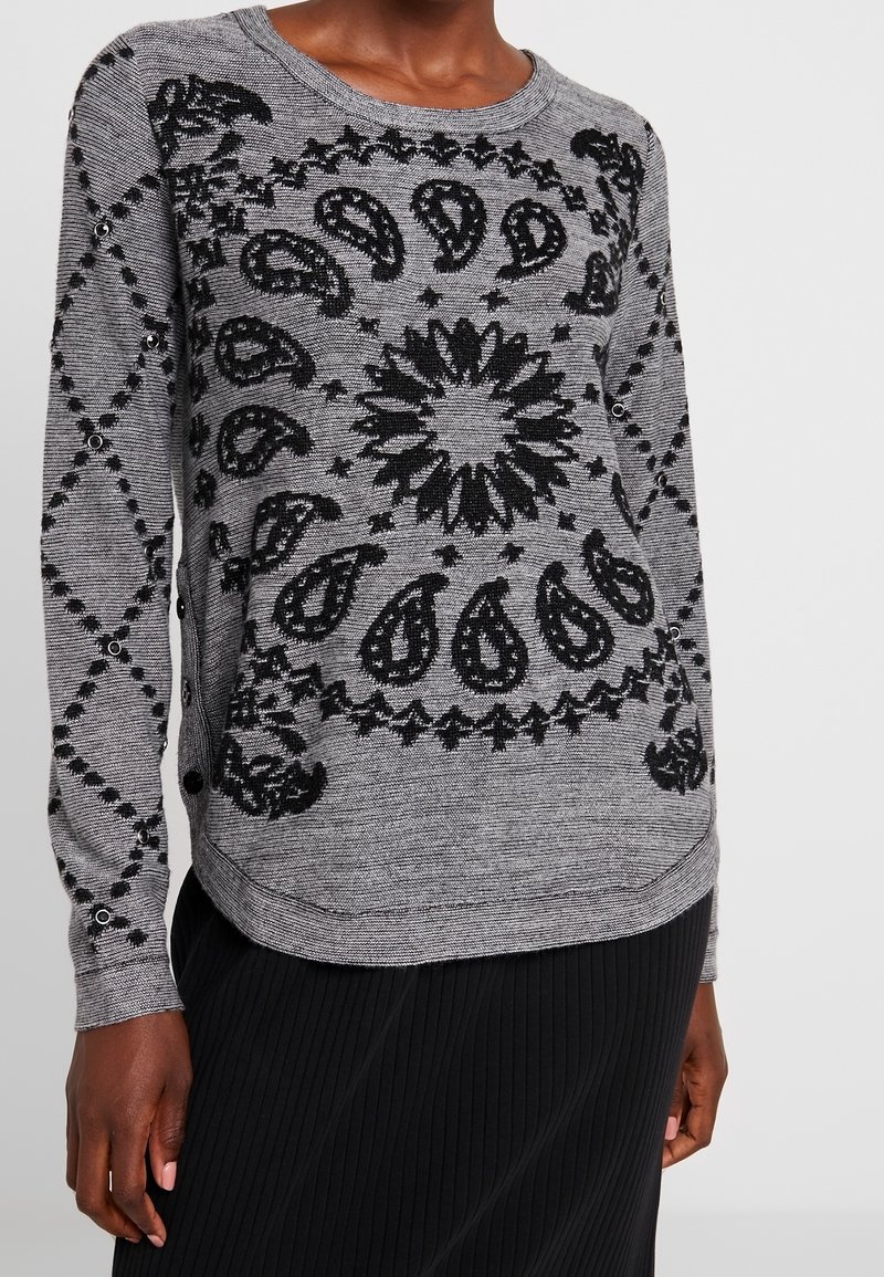 Pull en maille gris à manches longues avec motifs cachemire noirs et motifs floraux, porté avec une jupe plissée noire sur une personne à la peau foncée.