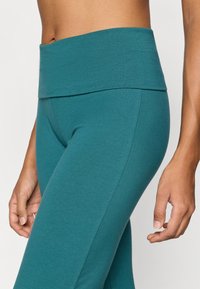 Τιρκουάζ κολάν με ψηλή μέση από μαλακό, ελαστικό ύφασμα. Διαθέτε ι ευρεία, διπλωμένη μέση και λεία, εφαρμοστή σιλουέτα.