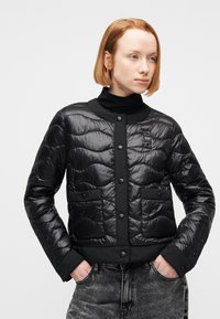 Νεαρό άτομο με κοντά κόκκινα μαλλιά που φοράει μαύρο μπουφάν με ραφές πάνω από μαύρο ζιβάγκο, με τα χέρια στις τσέπες, κοιτώντας προς το πλάι.