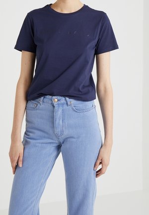 Osoba w granatowej koszulce z krótkim rękawem i wysokim stanem jasnoniebieskich dżinsów na tle jednolitym.