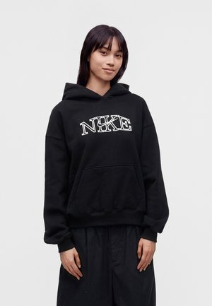 Giovane donna con capelli lisci e scuri indossa una felpa nera Nike con logo bianco, in piedi davanti a uno sfondo chiaro e semplice.