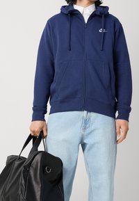 Person som bär en marinblå Nike-ziphoodie, ljusblå jeans och håller en stor svart läderduffelväska i vänster hand.