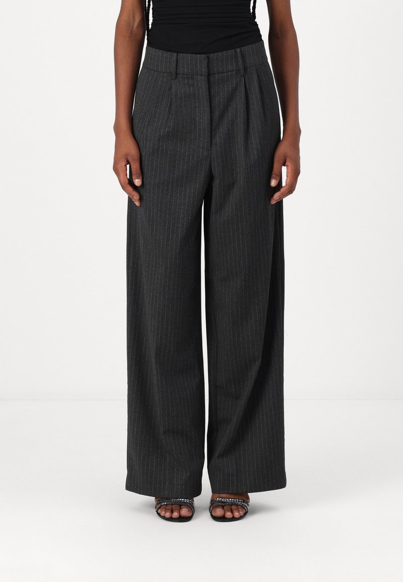 Pantaloni larghi a vita alta in tessuto grigio scuro con motivo a righe e pieghe frontali, abbinati a sandali neri con tacco aperto e cinturini.