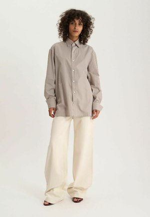 Femme aux cheveux bouclés portant une chemise boutonnée beige et blanche à rayures et un pantalon crème à jambes larges, debout devant un fond uni.