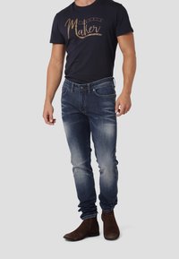 Jeans de mezclilla azul oscuro con corte slim, ligero desgaste y diseño clásico de cinco bolsillos. Combinados con una camiseta negra que presenta un gráfico en dorado.