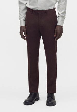 Homme portant un pantalon habillé marron foncé, des chaussures habillées noires et une chemise gris clair boutonnée sur un fond uni.