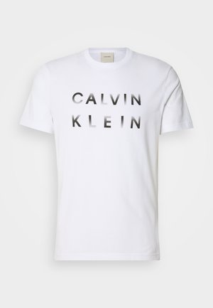Biela bavlnená tričko s krátkym rukávom a okrúhlym výstrihom, so značkou "CALVIN KLEIN" potlačenou tučnými čiernymi písmenami cez prednú časť.