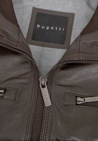 Braune Lederjacke mit einem Frontreißverschluss und zwei Brusttaschen, mit einer glatten Textur und einem grauen Innenfutter. Markenlabel sichtbar.