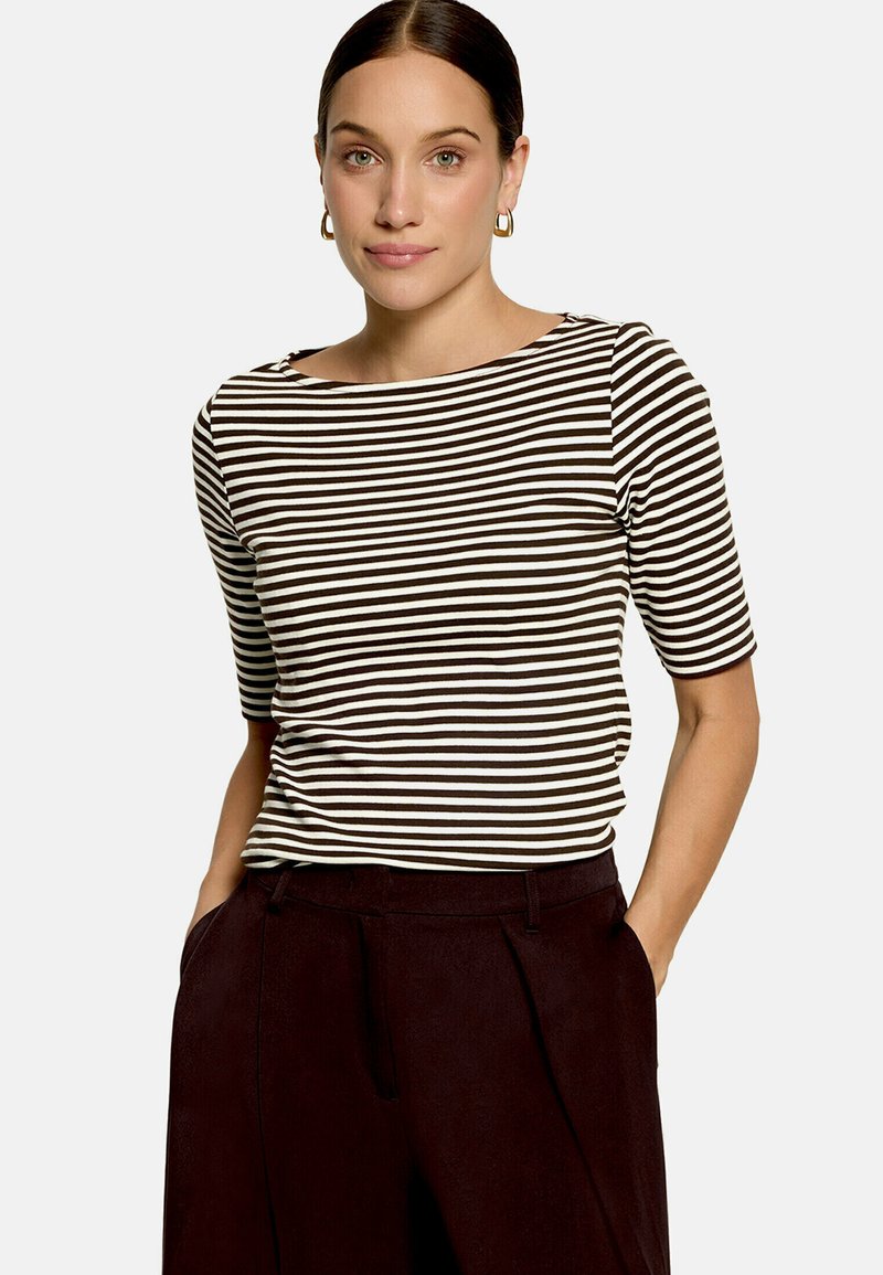 Femme aux cheveux foncés en chignon bas, portant des boucles d’oreilles créoles dorées, un t-shirt à manches courtes rayé noir et blanc, et un pantalon taille haute foncé, les mains dans les poches.