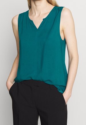Femme portant un chemisier sans manches bleu sarcelle avec un col en encoche et un pantalon noir, debout les mains dans les poches.