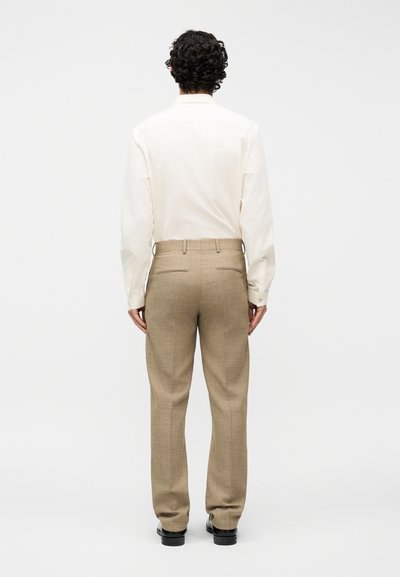 Άνδρας με σγουρά μαλλιά, φορώντας μπεζ παντελόνι φόρμας, λευκό πουκάμισο μακρύ μανίκι και μαύρα παπούτσια, στέκεται με την πλάτη σε απλό φόντο.