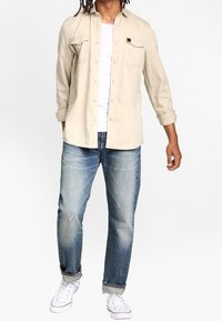 Helles beigefarbenes Hemd mit Knöpfen und zwei Brusttaschen, weißes T-Shirt, abgewetzte blaue Jeans und weiße Sneaker. Lässig, entspannte Passform.