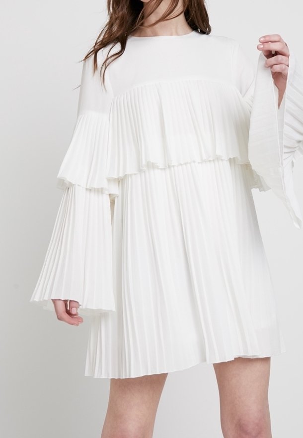 Robe plissée blanche avec des manches longues et évasées, et un design à étages. Tissu lisse avec un col rond. Tombe au-dessus du genou.