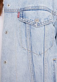 Jasnoniebieska kurtka dżinsowa z metalowymi zatrzaskami i kieszonką z klapką, na której znajduje się czerwona metka Levi's na piersi.