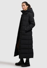 Cămașă puffer lungă, de culoare neagră, cu guler înalt, închidere cu fermoar frontală și buzunare laterale. Prezintă o textură matlasată și o siluetă dreaptă.