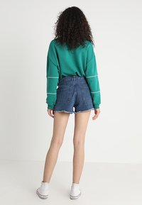 Sudadera verde de gran tamaño con líneas blancas, combinada con shorts de denim deshilachados de tiro alto. La modelo usa zapatillas blancas.