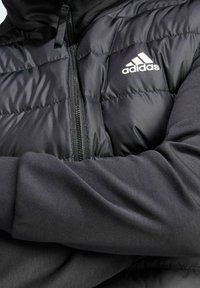 Czarna kamizelka puchowa o pikowanej teksturze i srebrnym zamku błyskawicznym, z haftowanym białym logo na piersi, noszona na czarnej bluzie z długim rękawem.