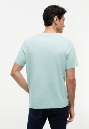 Άνδρας με σκούρα μαλλιά που φοράει ένα ανοιχτό μπλε κοντομάνικο μπλουζάκι και σκούρα τζιν, γυρισμένος με την πλάτη σε ένα απλό λευκό φόντο.