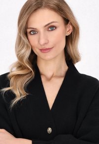 Cardigan nero con tessuto a trama, scollo a V profondo e un prominente dettaglio con bottone dorato. La modella mette in mostra il design con capelli morbidi e mossi.