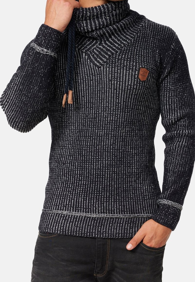 Homme portant un pull en maille côtelée noir et blanc avec col montant, cordons et détail en cuir sur la poitrine, main dans la poche.