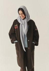 Barna gyapjúkabát virágmintás hímzéssel, világoskék pulóver rétegezve alatta, világosszürke kötött sállal párosítva. Két elülső zseb látható.