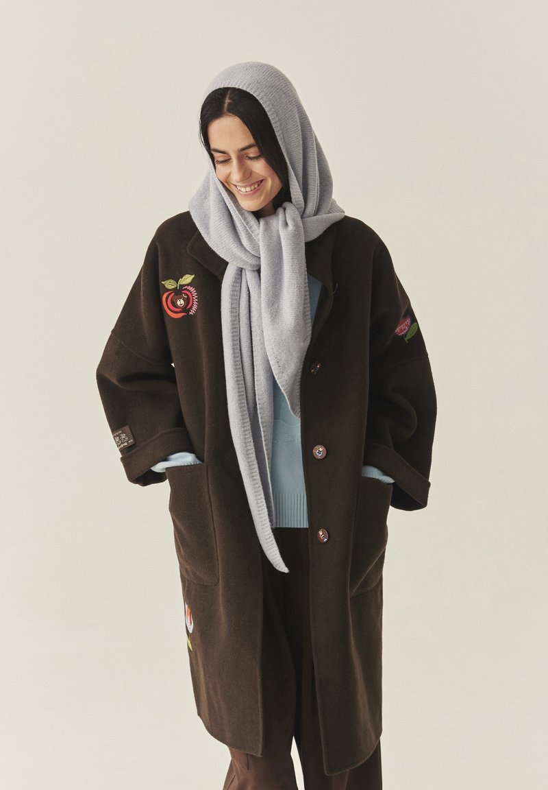 Barna gyapjúkabát virágmintás hímzéssel, világoskék pulóver rétegezve alatta, világosszürke kötött sállal párosítva. Két elülső zseb látható.