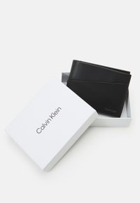 Black leather wallet with textured finish, displayed in a white box labeled "Calvin Klein." Features a simple, modern design.