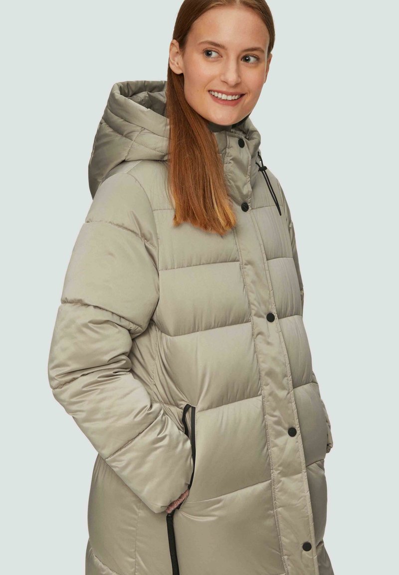 Helle beigefarbene Daunenjacke mit Kapuze, ausgestattet mit gestepptem Material, Druckknöpfen und Seitentaschen mit schwarzen Kordelzügen.