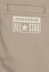 Beige tyglabel med "CONVERSE ALL STAR" i fet svart text och en stjärnsymbol, sytt på en texturerad canvasbakgrund.
