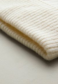 Valge kootud müts, valmistatud pehmest ja tekstuurilisest villast, millel on soonikkoes disain ja allapoole volditud ääred.