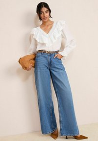 Blusa bianca con colletto arricciato e bordo in pizzo, abbinata a jeans a gamba larga blu e tacchi color carne. In mano tiene una pochette in suede marrone.