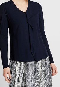 Marinblus med långa ärmar och vikdesign samt v-ringning. Matchad med en silver- och svart mönstrad kjol med orm-mönster, som visar lager av texturer.