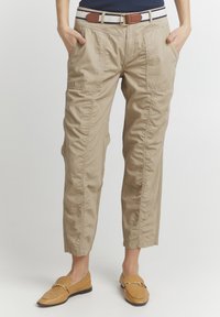 Pantalons cargo beiges avec poches latérales, retroussés à la cheville, associés à des mocassins marron clair. Le tissu semble léger et texturé.