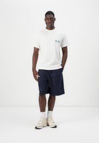 Camiseta de algodón blanca con un gráfico azul en el bolsillo, combinada con pantalones cortos azul marino hasta la rodilla. Zapatillas beige con calcetines gris claro.