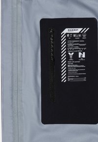 Grauer Stoff mit einem schwarzen Patch, der Sicherheitshinweise enthält. Der Patch umfasst einen Reißverschluss und auffällige weiße Symbole und Texte für Notfallsignale.