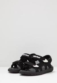 Czarne sandały na otwartym palcu z regulowanymi paskami na rzep, gumową podeszwą i białymi akcentami. Teksturowana wkładka dla lepszej przyczepności i komfortu.