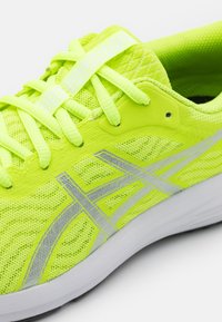 Neon žlutá sportovní bota s síťovaným svrškem, reflektivními stříbrnými akcenty a plochými tkaničkami. Obsahuje polstrovanou podrážku a mírně strukturovaný detail.