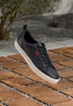 Sort lavtop sneaker med hvid sål og rød "HUGO" tag, vist over firkantede træfliser med grus imellem, udendørs med træ.