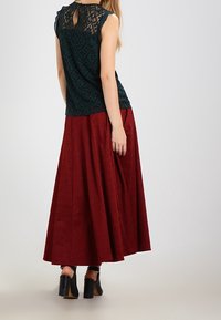 Blusa de encaje verde oscuro con textura estampada, combinada con una falda plisada burdeos hasta el tobillo. Zapatos de tacón negros completan el look.