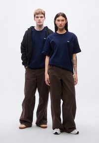 Ναυτικά βαμβακερά μπλουζάκια, φαρδιά γραμμή, με λεπτομέρεια λογότυπου. Καφέ παντελόνια φαρδιάς γραμμής, casual σχεδιασμός. Μαύρο μπουφάν με φερμουάρ και κουκούλα, μαζί με σκούρα αθλητικά παπούτσια.