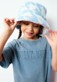 Ljusblå bucket-hatt med vitt stjärnmönster, mjukt fleecematerial. Blå t-shirt med "NEW YORK" broderat i mörkare blå tråd.