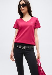 Γυναίκα με σκούρα γυαλιά ηλίου, ροζ μπλουζάκι με V λαιμόκοψη, μαύρο παντελόνι με ζώνη με τρουκς, κρατώντας μια ροζ τσάντα χειρός μπροστά σε απλό φόντο.