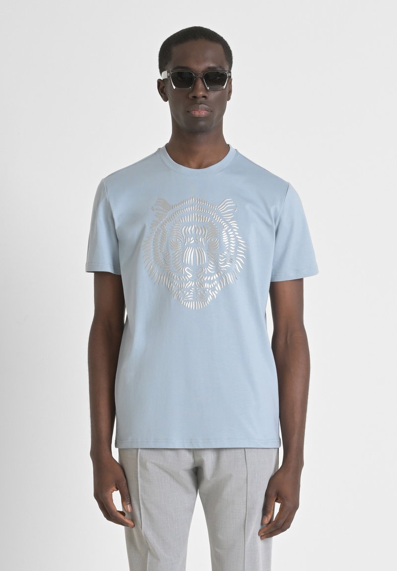 Camiseta de manga corta azul claro con un gráfico de tigre plateado en el frente, hecha de tela suave. El modelo usa gafas de sol negras.