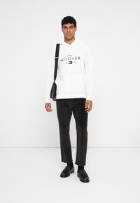 Hvit hettegenser med "Tommy HILFIGER"-logo, svarte corduroy-bukser, svarte sko og en liten skulderveske. Enkelt design og nøytrale farger.