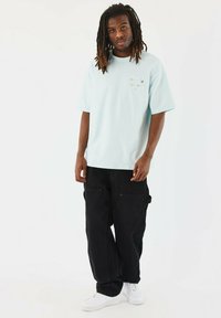Camiseta azul claro de gran tamaño con mangas cortas y un pequeño detalle gráfico, combinada con pantalones cargo negros y zapatillas blancas.