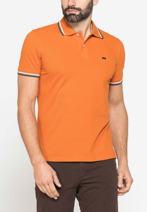 Uomo che indossa una polo arancione a maniche corte con collo e polsini a righe, abbinata a pantaloni marrone scuro, in piedi davanti a uno sfondo bianco.