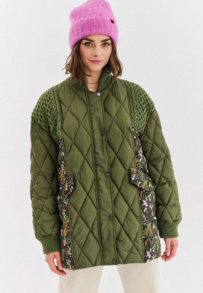 Zelená prošívaná bunda s barevným vzorem na boku a pletenými zelenými doplňky. Má velké našité kapsy, zapínání na knoflíky a žebrované manžety. Na hlavě růžová čepice.