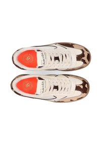 Baskets blanches avec accents en imprimé vache, surface texturée, semelle intérieure orange et lacets blancs. Présente une étiquette de logo sur la languette et des lignes noires contrastantes.