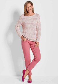 Strickpullover in Pink mit strukturiertem Muster, kombiniert mit slim-fit hellrosa Hosen und pinken spitzen Ballerinas. Das Model posiert mit Händen in den Hüften.